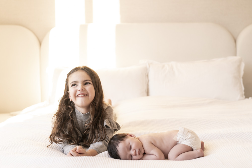 happy girl with long hair lying on her belly next to sleeping newborn in diapers
