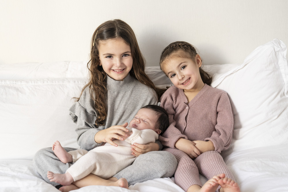 two smiling toddlers sitting on a bed while the one on the left is holding a sleeping newborn