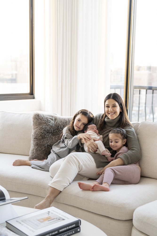 mother holding her sleeping newborn sitting in the middle of two toddlers on a couch in front of a window all are smiling at the camera