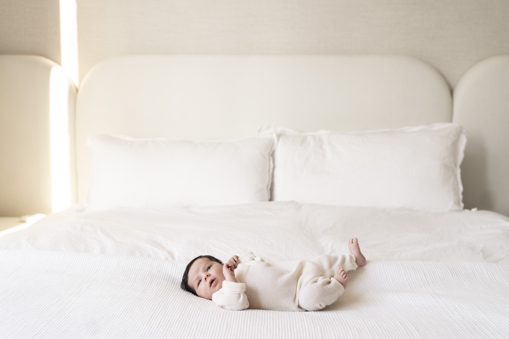 newborn wearing a beige onesie lying on a bed with white sheets looking at the camera
