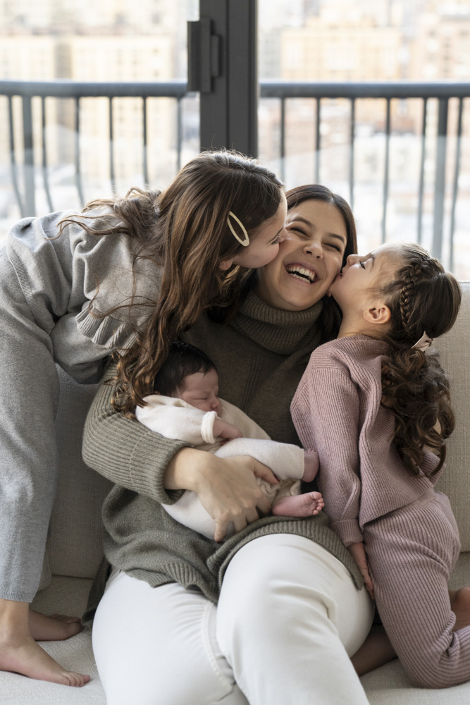 smiling mother sitting on the couch holding her sleeping newborn her two toddlers sitting next to her one on each side and are kissing her cheek