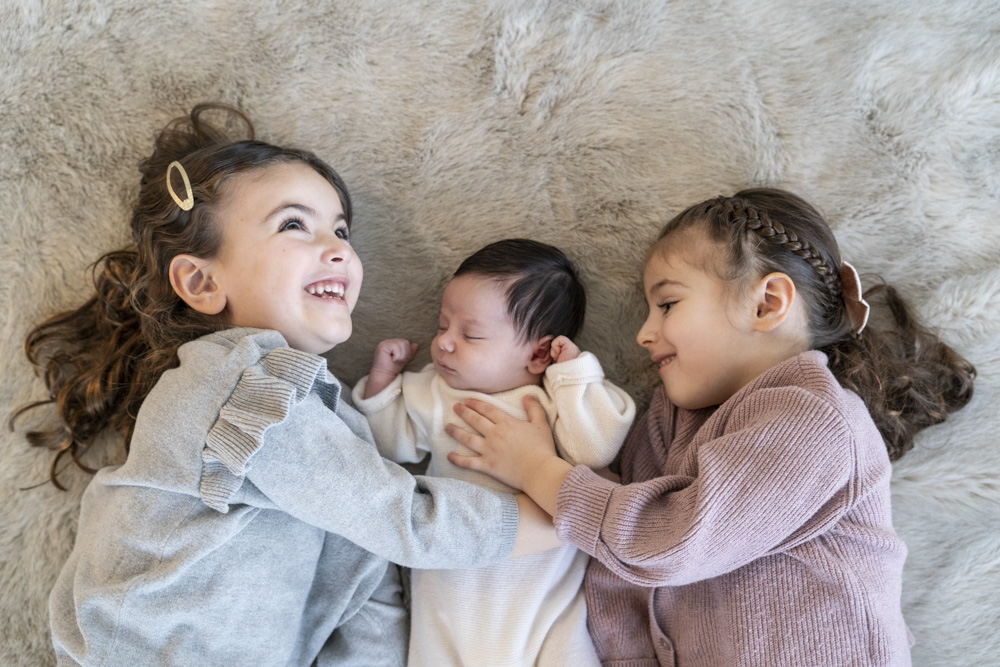 sleeping newborn lying in the middle of two smiling toddlers both are touching the bab