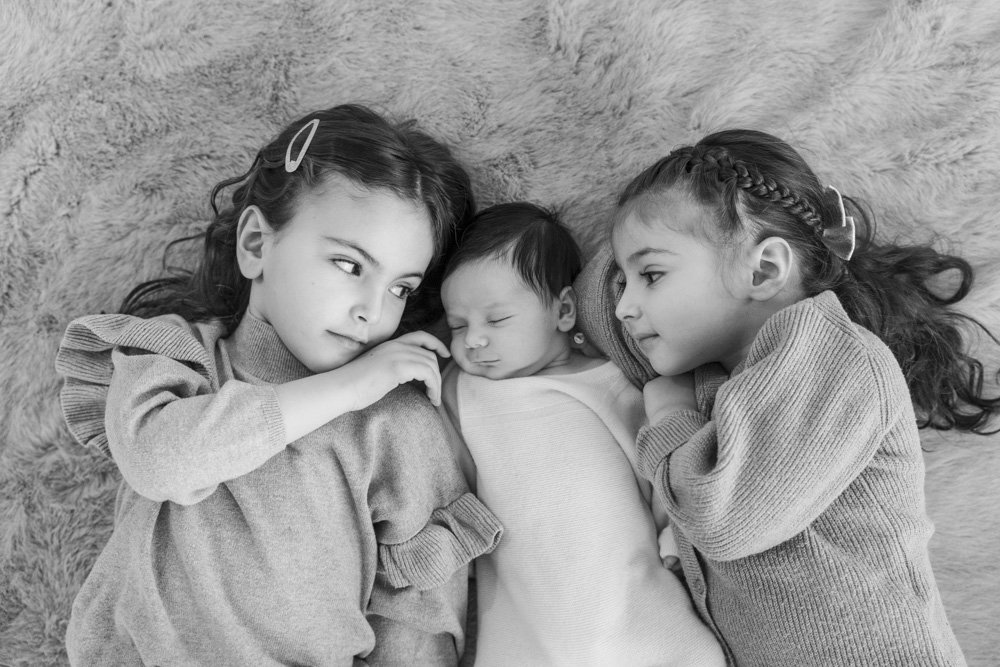 black and white photo of a sleeping newborn lying in the middle of two toddlers both are smiling at the baby