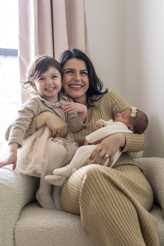 mother sitting in a chair holding her sleeping newborn in her left arm and her toddler in her right arme both are smiling at the camera