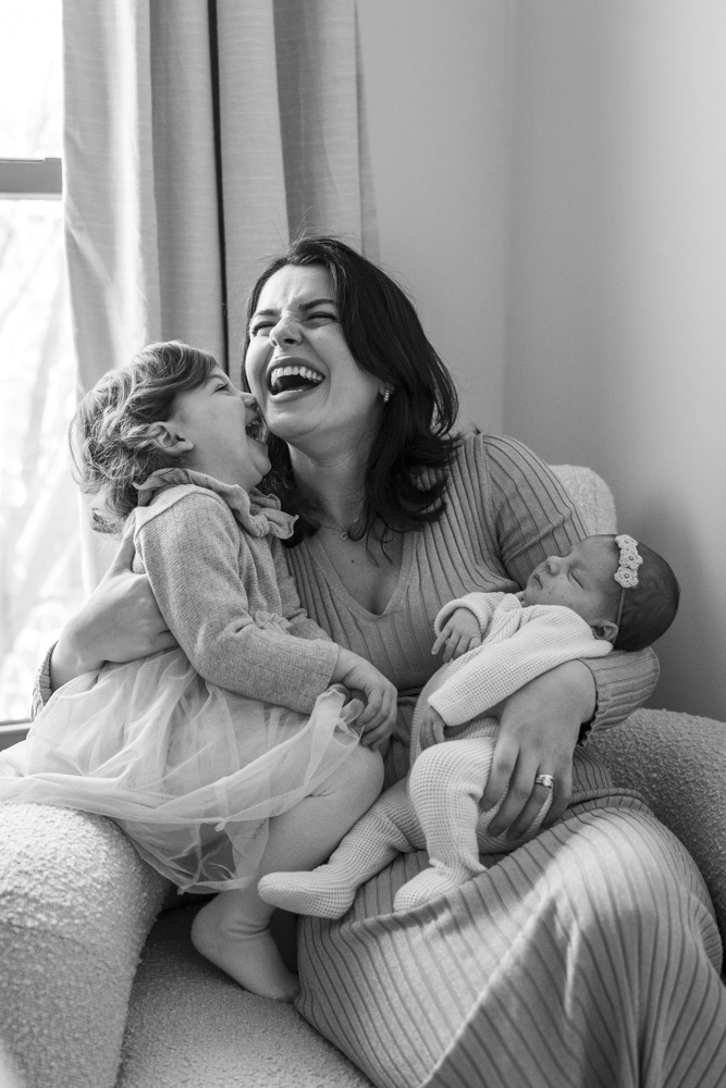 black and white photo of mother sitting in a chair holding her sleeping newborn in her left arm and her toddler in her right arme both are laughing