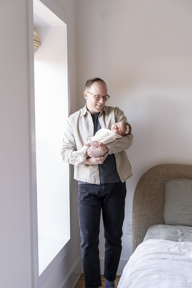 father standing next to a window holding his sleeping newborn in his arms smiling at the baby