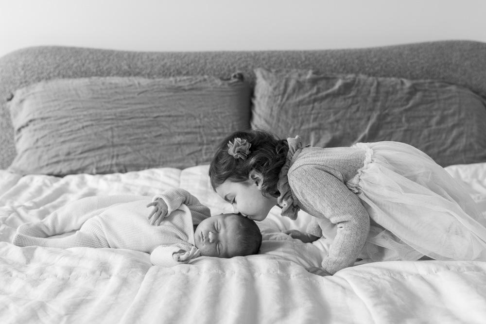 black and white of a sleeping newborn lying on the bed wearing a onesie the big sister is kissing the baby's head