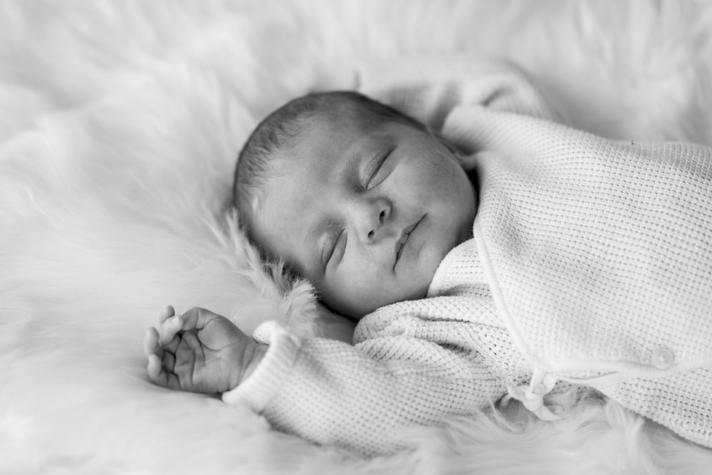 black and white photo of a sleeping newborn wearing a white onesie laying on a white fuzzy blanket