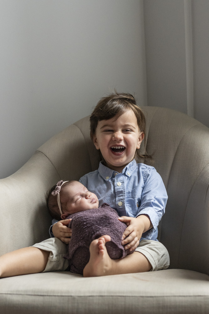 toddler sitting in a chair holding the sleeping newborn laughing at the camera