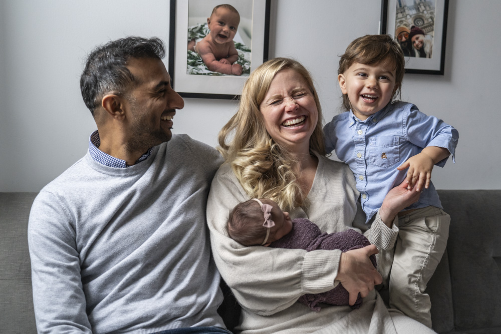 happy family of four sitting on the couch mother is sitting in the middle holding her newborn laughing father is sitting on one side laughing at her and toddler is standing in the other side smiling at the camera