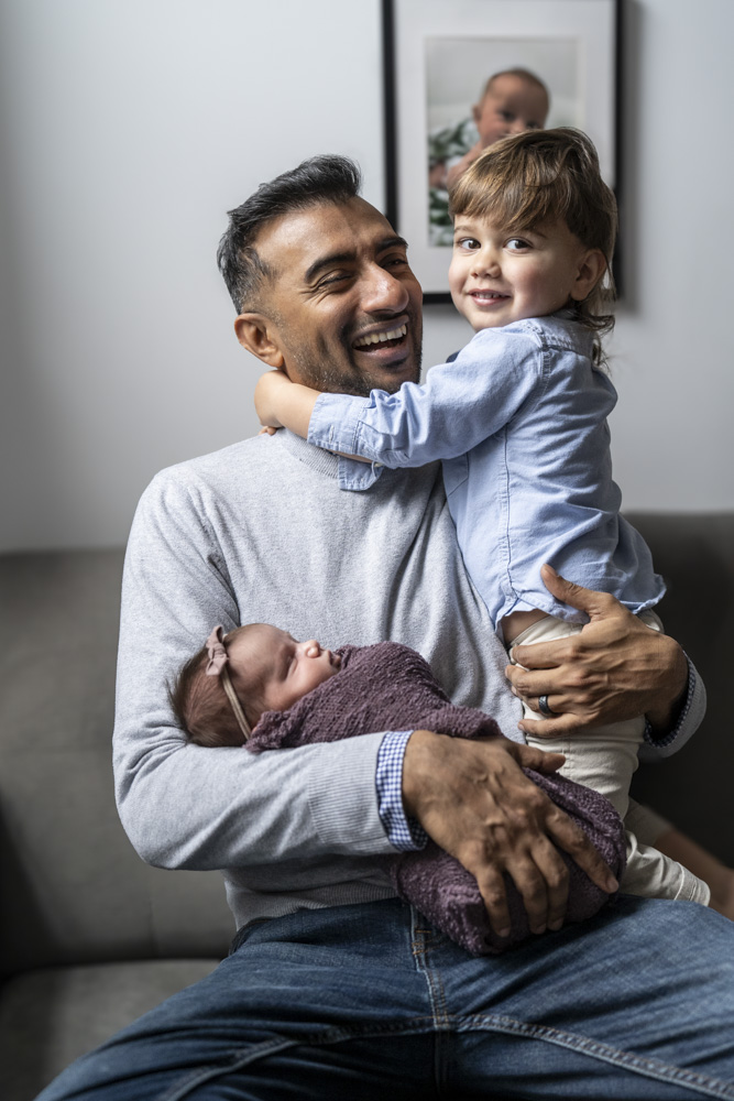 father is holding his newborn in his right arm and holding his toddler with his other arm toddler wrapped his arms around his father's neck smiling at the camera father is laughing at the toddler