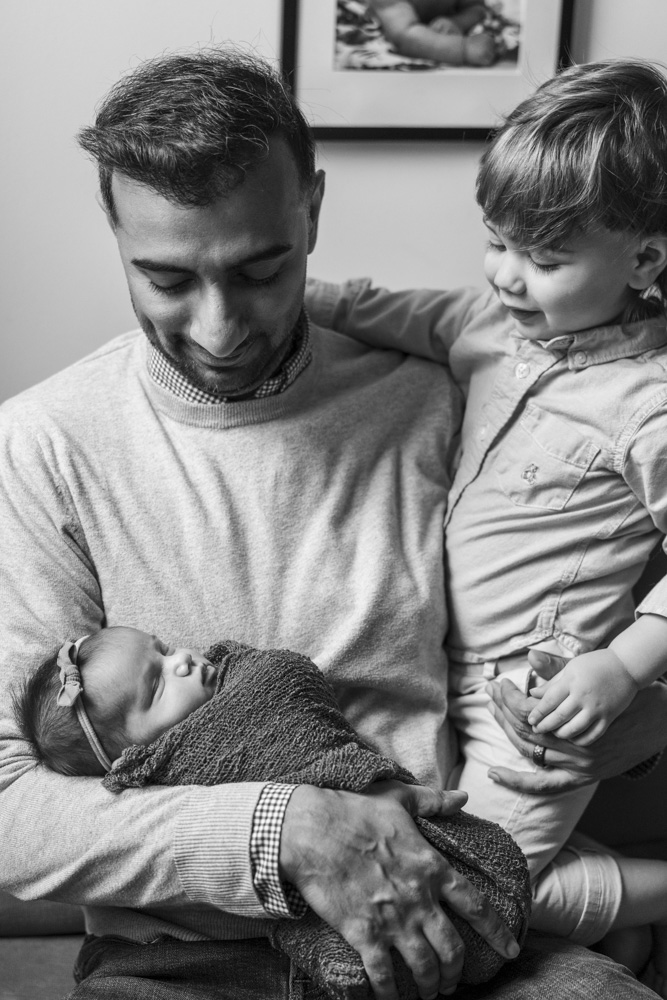 black and white photo of a father holding his sleeping newborn while wrapping his other arm around his toddler both smiling at the baby