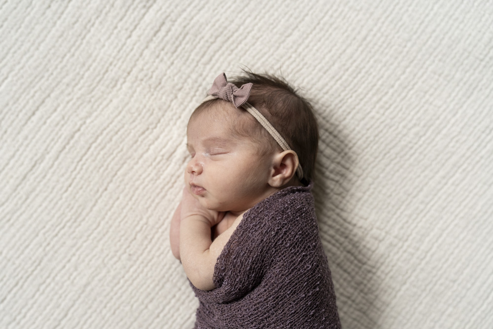 close up of newborn wearing a headband wrapped up in a brown blanket sleeping on its side with hands tucked under the chin lying on a beige blanket