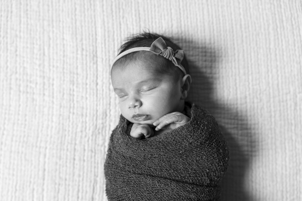 black and white photo of a sleeping newborn wearing a headband swaddled in a blanket with hands sticking out