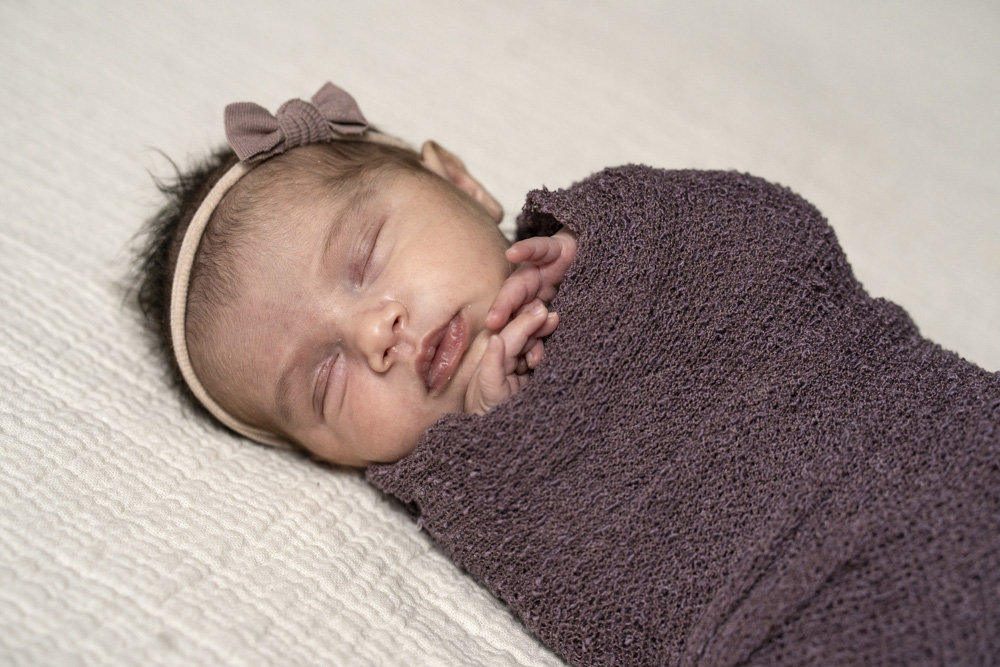 close up of a sleeping newborn wearing a headband swaddled in a brown blanket lying on a beige blanket with hands sticking out
