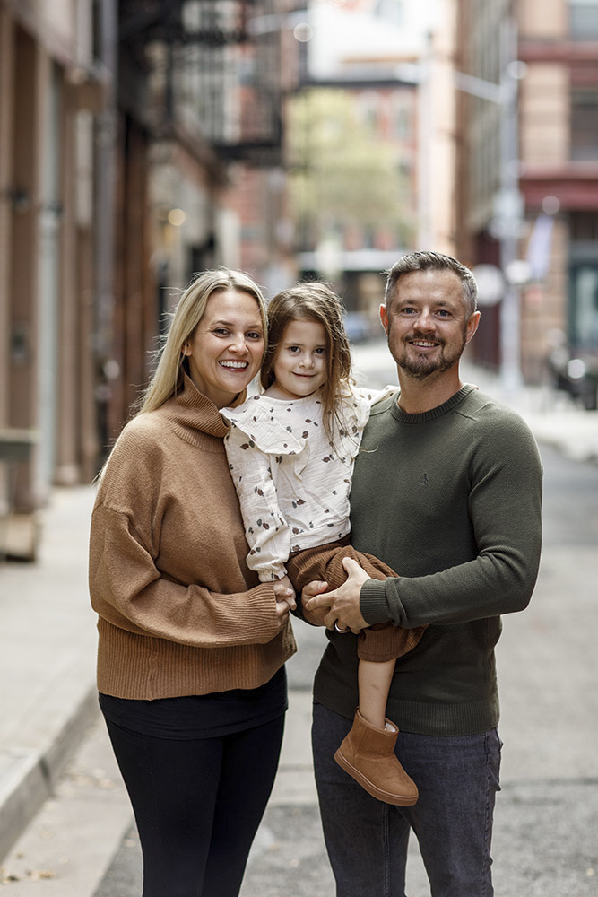family of three in the streets of New York smiling at the camera while father is holding his daughter