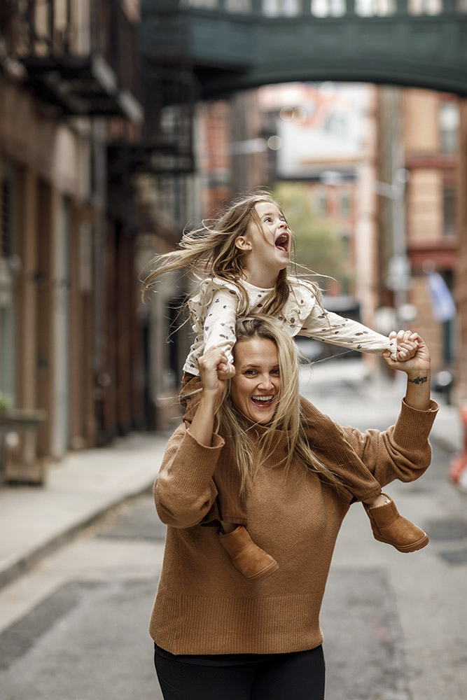 happy little girl is sitting on her mother's shoulders holding her hands while mom is swirling her around both laughing