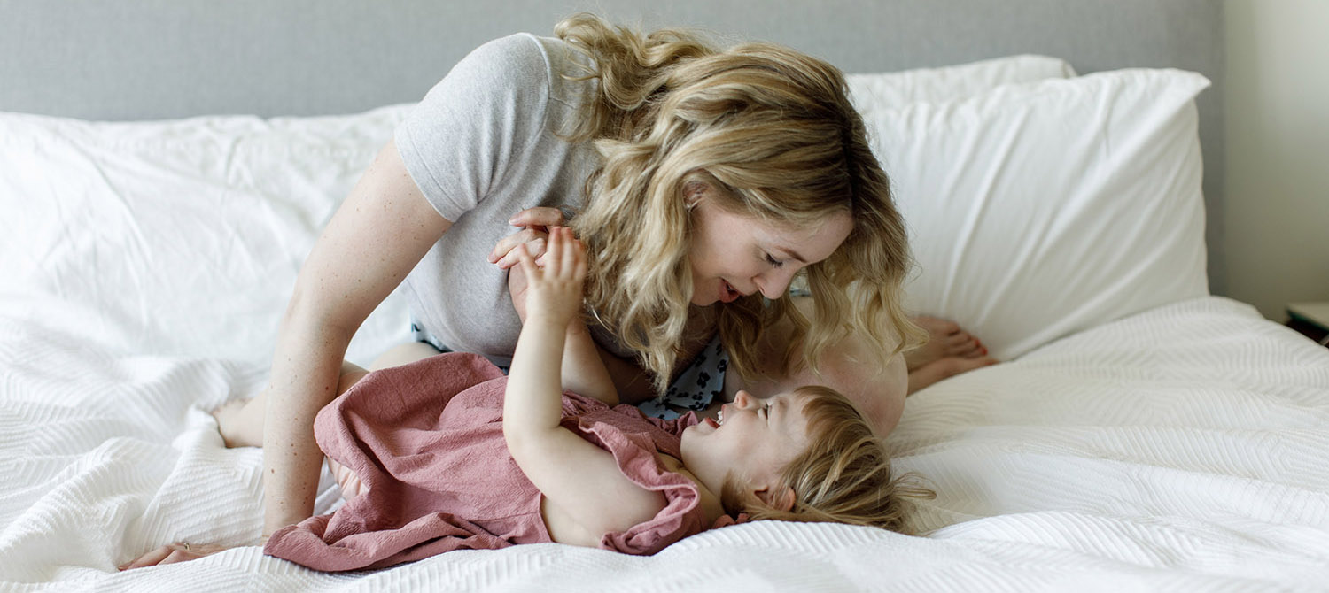 mother is goofing around with her daughter while daughter is laying on her back on a big bed with white sheets