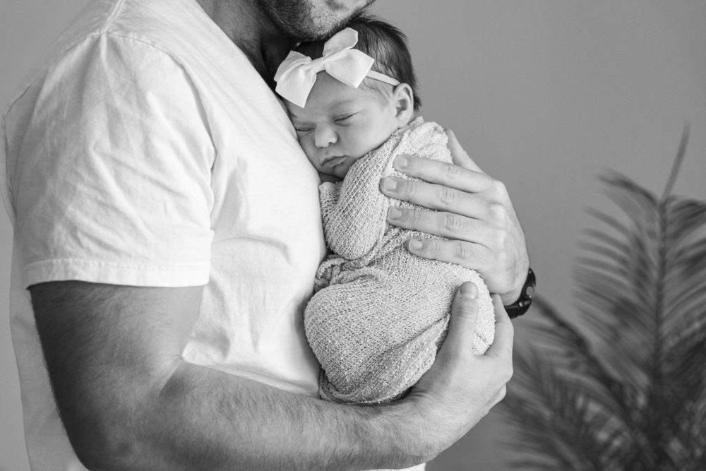 black and white photo of father holding the newborn close to his chest newborn is swaddled in a blanket and wearing a headband with a bow
