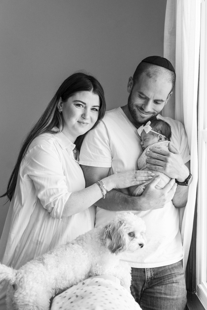 black and white photo of a family of three with their dog standing by the window father is holding the newborn