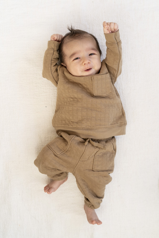 newborn in a brown outfit laying on a bed with white sheets smiling at the camera with arms up