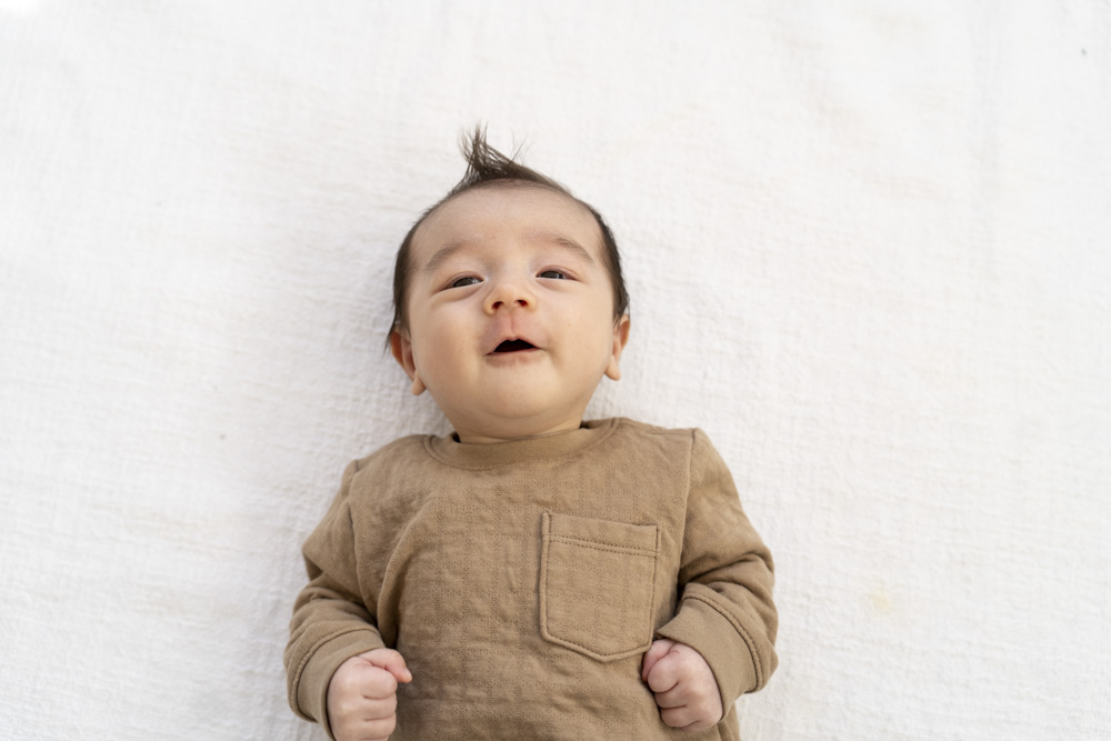 newborn wearing a brown shirt lying on a bed with white sheets looking at the camera