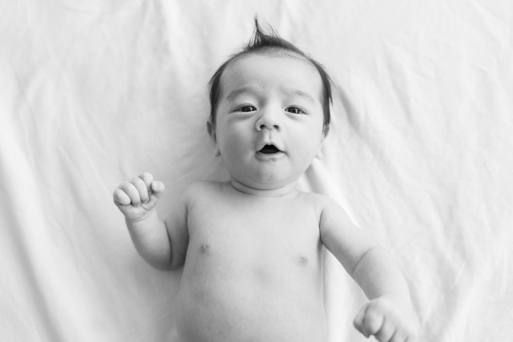 black and white photo of a newborn with dark hair lying on a white blanket looking at the camera
