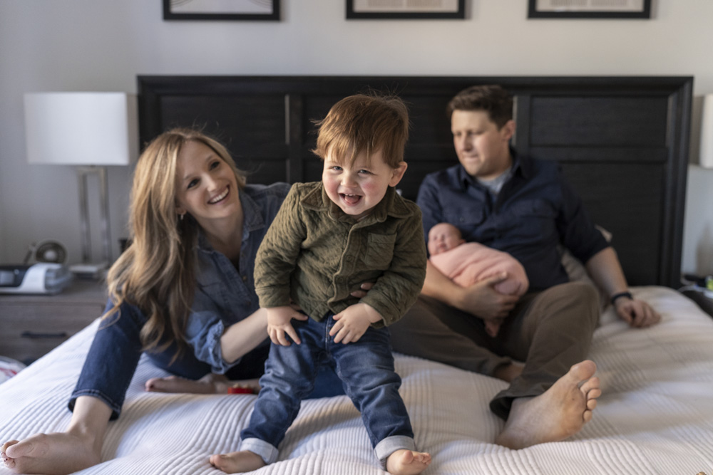family of four sitting on a bed toddler is laughing at the camera father is holding the newborn