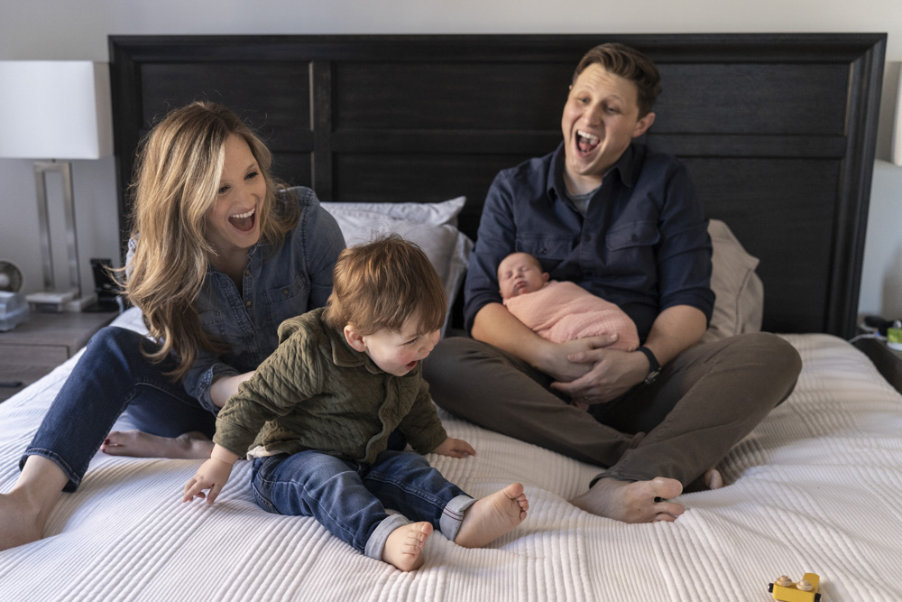 family of four sitting on a bed laughing and having fun father is holding the newborn
