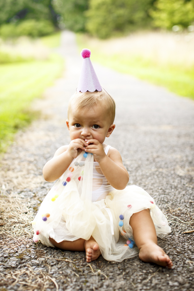 toddler wearing a birthday hat sitting on the ground while chewing on her dress looking at the camera