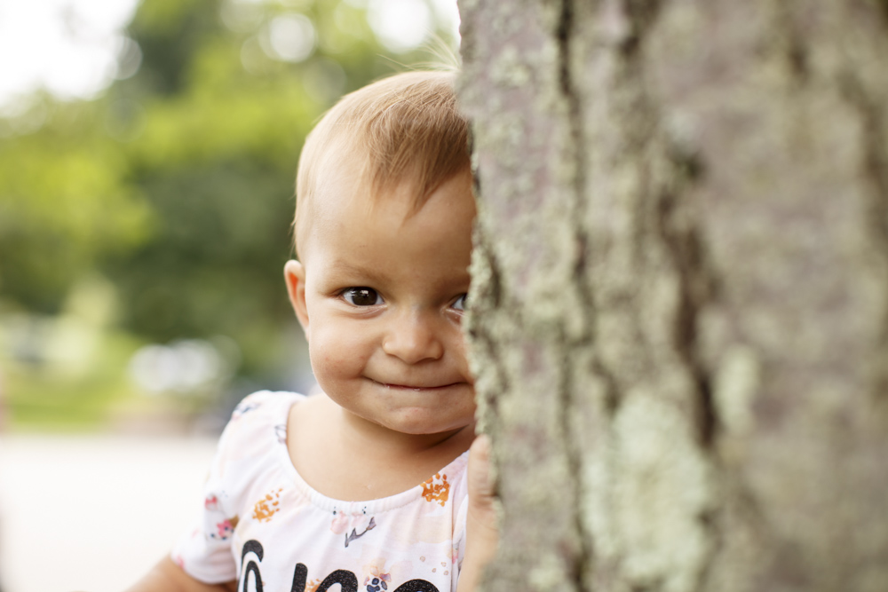 close up of a little girl peeking from behind a tree smiling at the camera