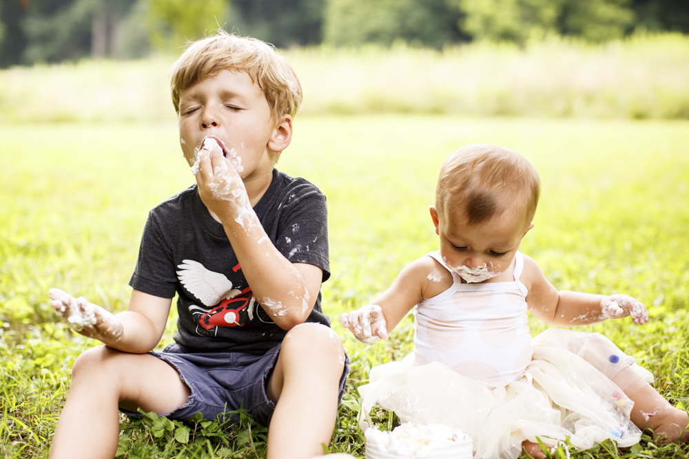 toddler and older brother are sitting in the grass eating cake hands and mouths are covered in cake