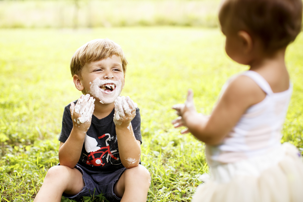 little boy is sitting in the grass his hands and mouth is covered in cake while toddler is watching him