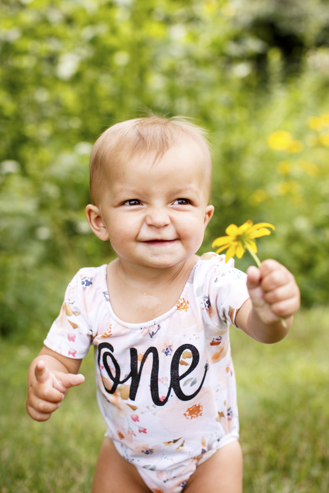 beautiful little birthday girl in a park wearing a onesie and holding a yellow little flower is standing outside with green trees in the background smiling at the camera