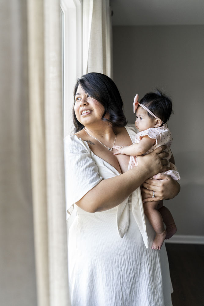 Baby Photography, mother in a white dress is standing next to a window holding her baby girl, baby wearing a beautiful dress and a bow headband