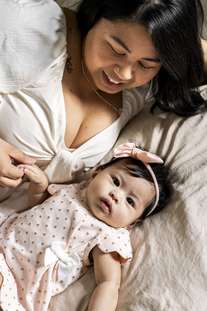 Baby Photography, mother and her baby girl lying next to each other on the bed, baby wearing a beautiful dress and a bow headband looking at the camera while mother is holding her hand and looking at her