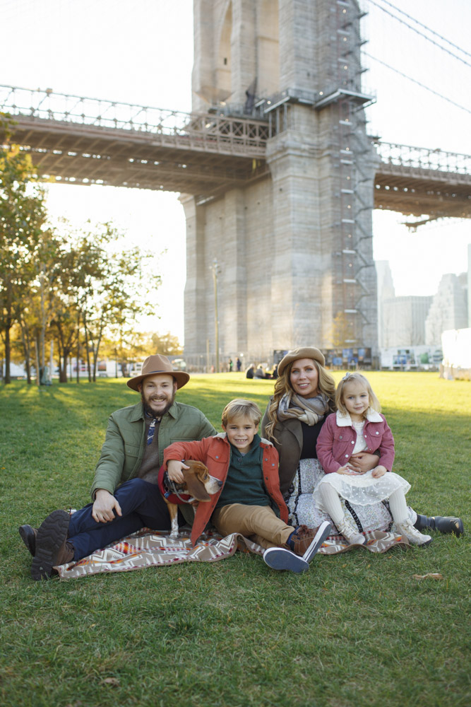 Family Session, family of four and their dog are sitting next to each other in the grass, the background shows the Brooklyn Bridge