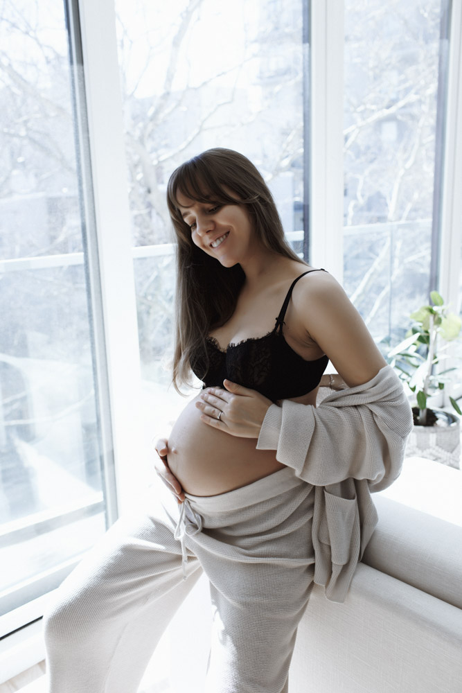 Maternity Session, pregnant woman standing in front of a big window holding her belly smiling at it