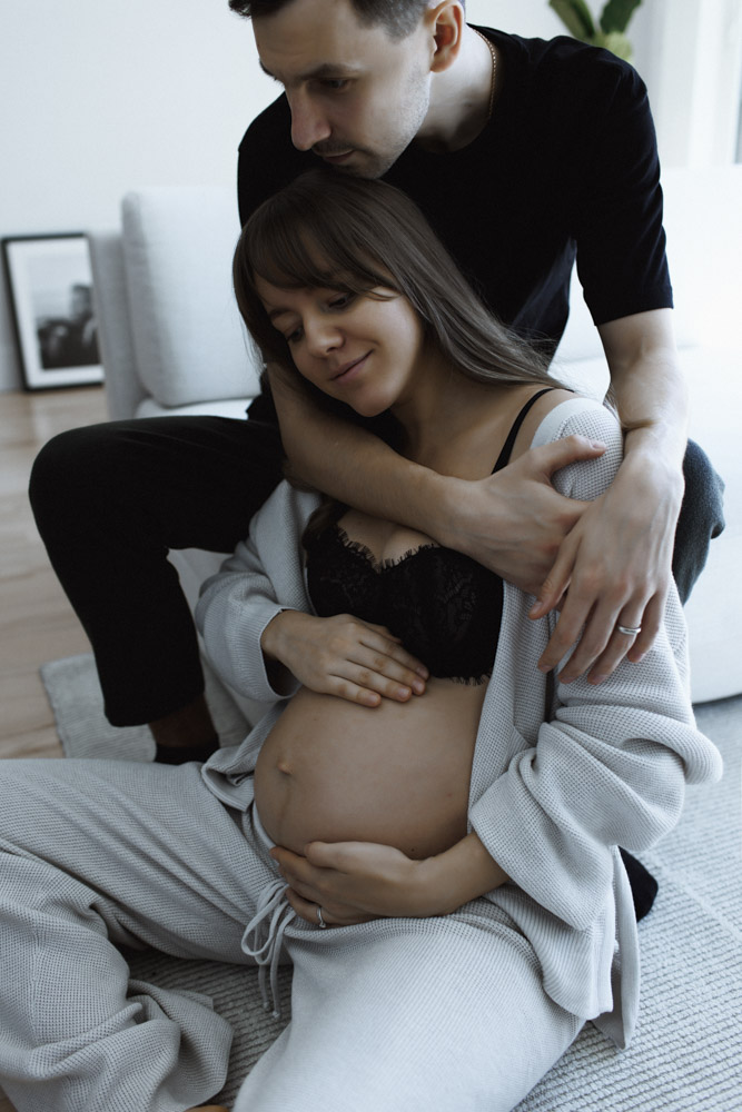 Maternity Session, pregnant woman is sitting on the floor touching her belly while partner is hugging her from behind