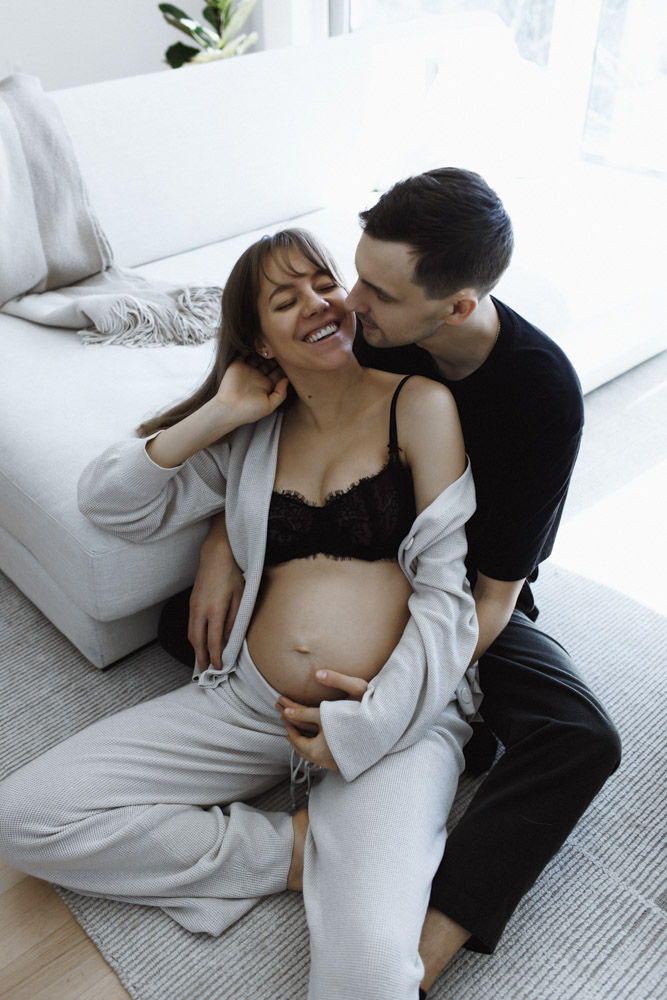 Maternity Session, pregnant woman is sitting on the floor leaning against her partner smiling while he is touching her belly
