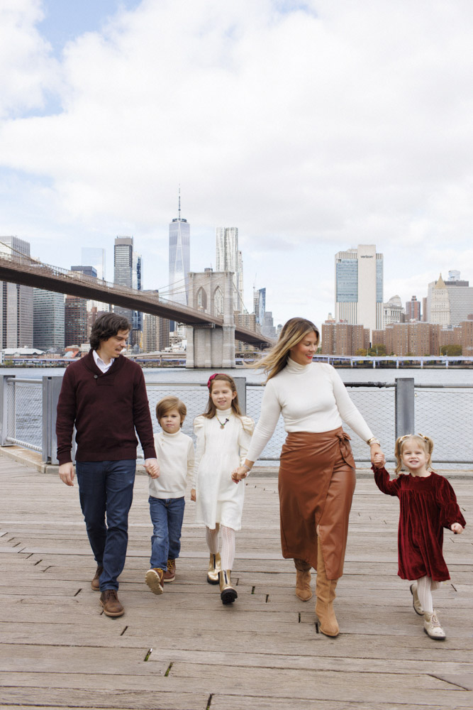 Family Session, family of five are walking on a pier holding hands, the background is the Brooklyn Bridge