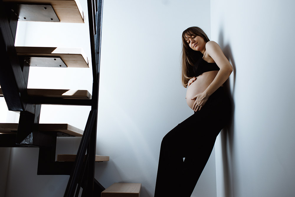 Maternity Session, pregnant woman holding her belly dressed in black is leaning agains a wall looking down