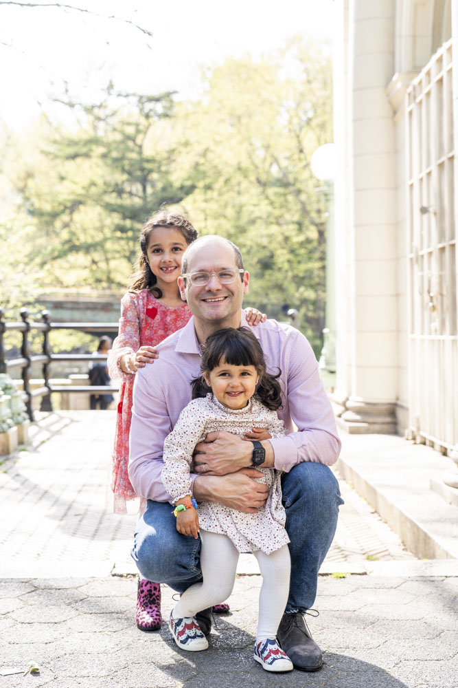 Spring Mini Session, father and his two daughters standing behind each other next to a building in a park smiling at the camera