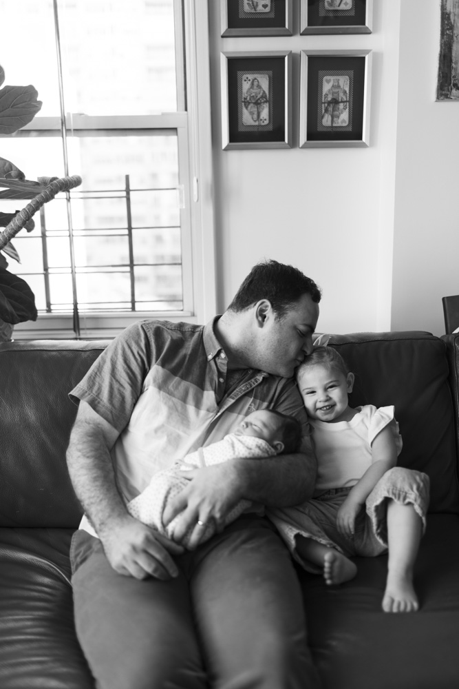 Family Photography, black and white photo of father sitting on the couch holding the sleeping newborn baby and kissing his toddler on her head while she is sitting next to him smiling at the camera