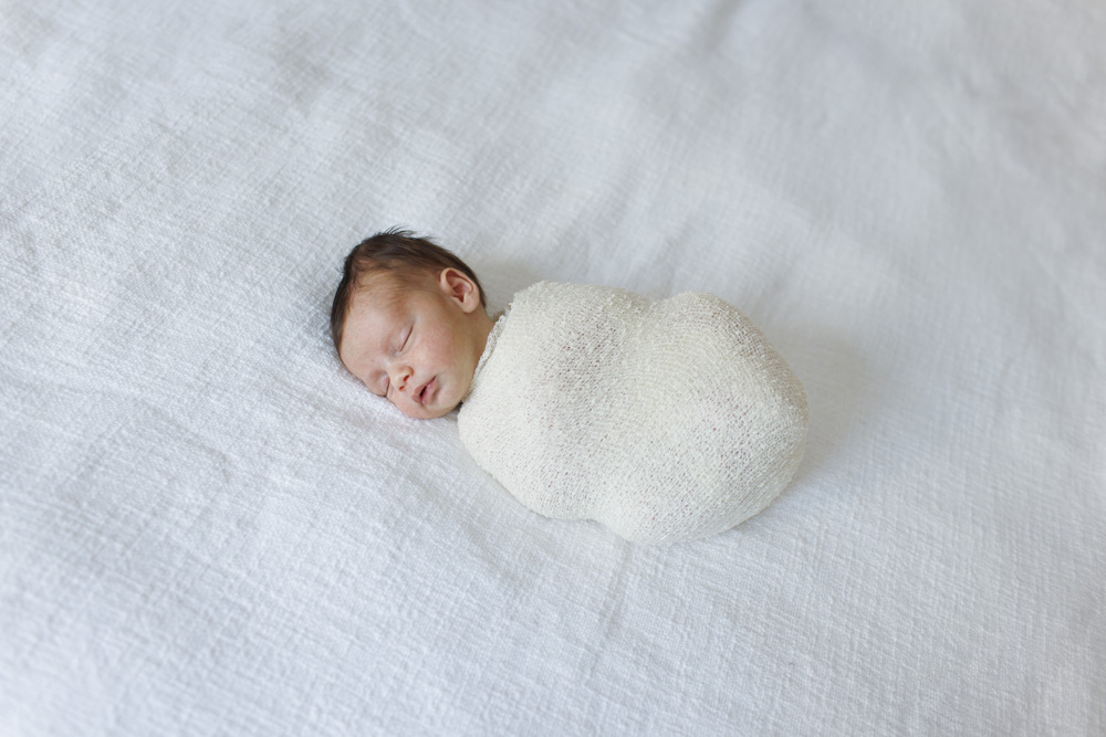 Baby Photography, newborn lying on a white sheet swaddled in a beige blanket