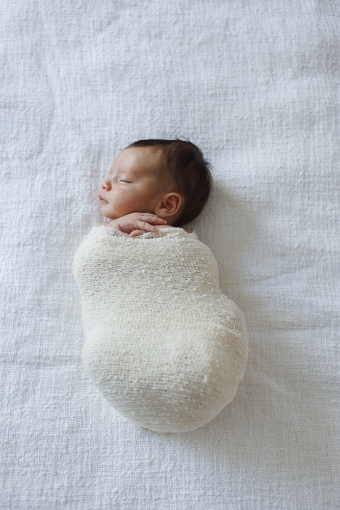 Baby Photography, newborn lying on a white sheet swaddled in a beige blanket with its hands sticking out eyes closed