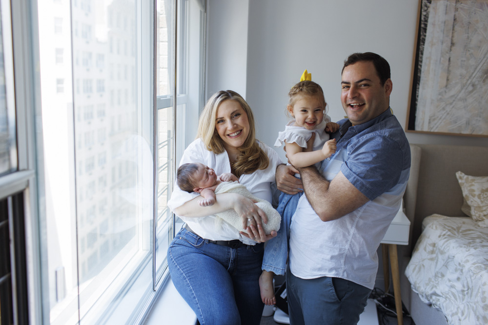Family Photos, Family of four standing next to a big window, mom is holding her sleeping newborn baby and dad is holding the big sister, all are laughing at the camera