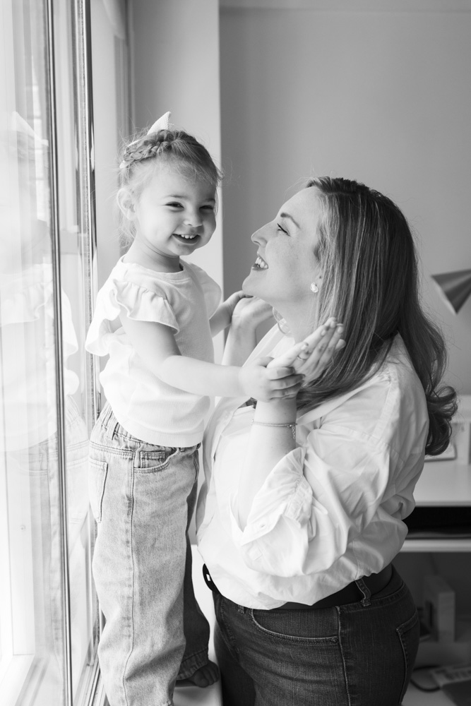 Family Session, beautiful black and white photo of mother and daughter, little girl is standing on the window sill while mom is standing in front of her holding her hands, both are laughing
