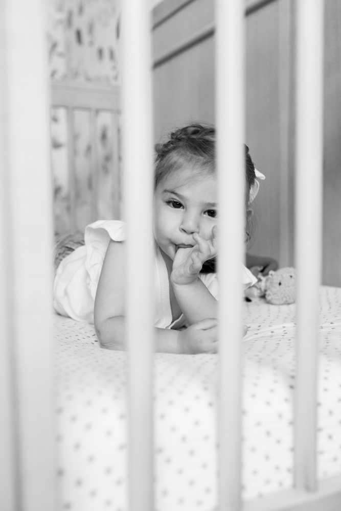 Family Photography, black and white photo of a little girl lying on her belly in the crib of her newborn sibling