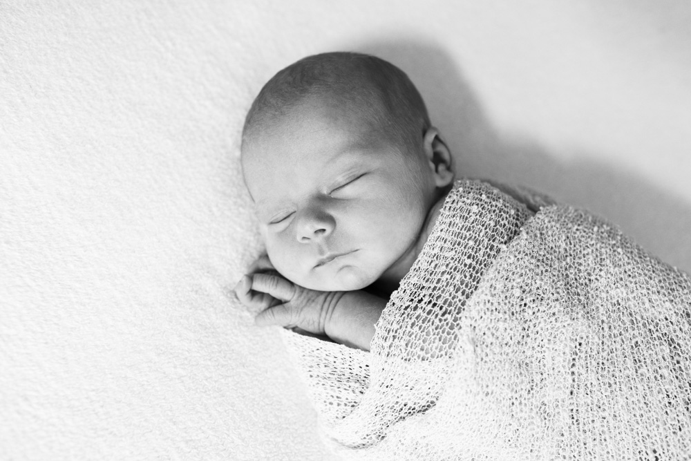 Baby Photography, black and white photo of a newborn swaddled in a light blanket with its hands sticking out eyes closed